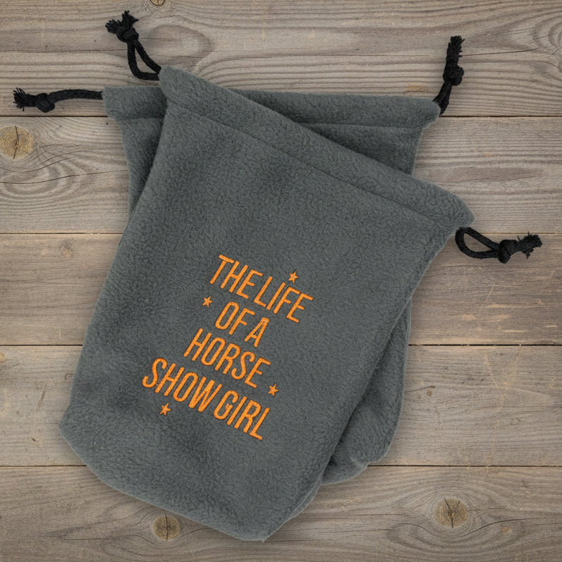 Gray stirrup covers with "The Life of a Horse Show Girl" embroidered in orange thread on a wooden surface